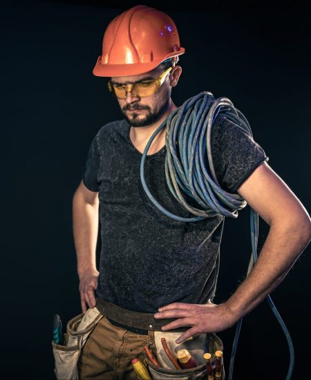 Male electrician in a protective helmet with an electric cable on a black background, copy space.